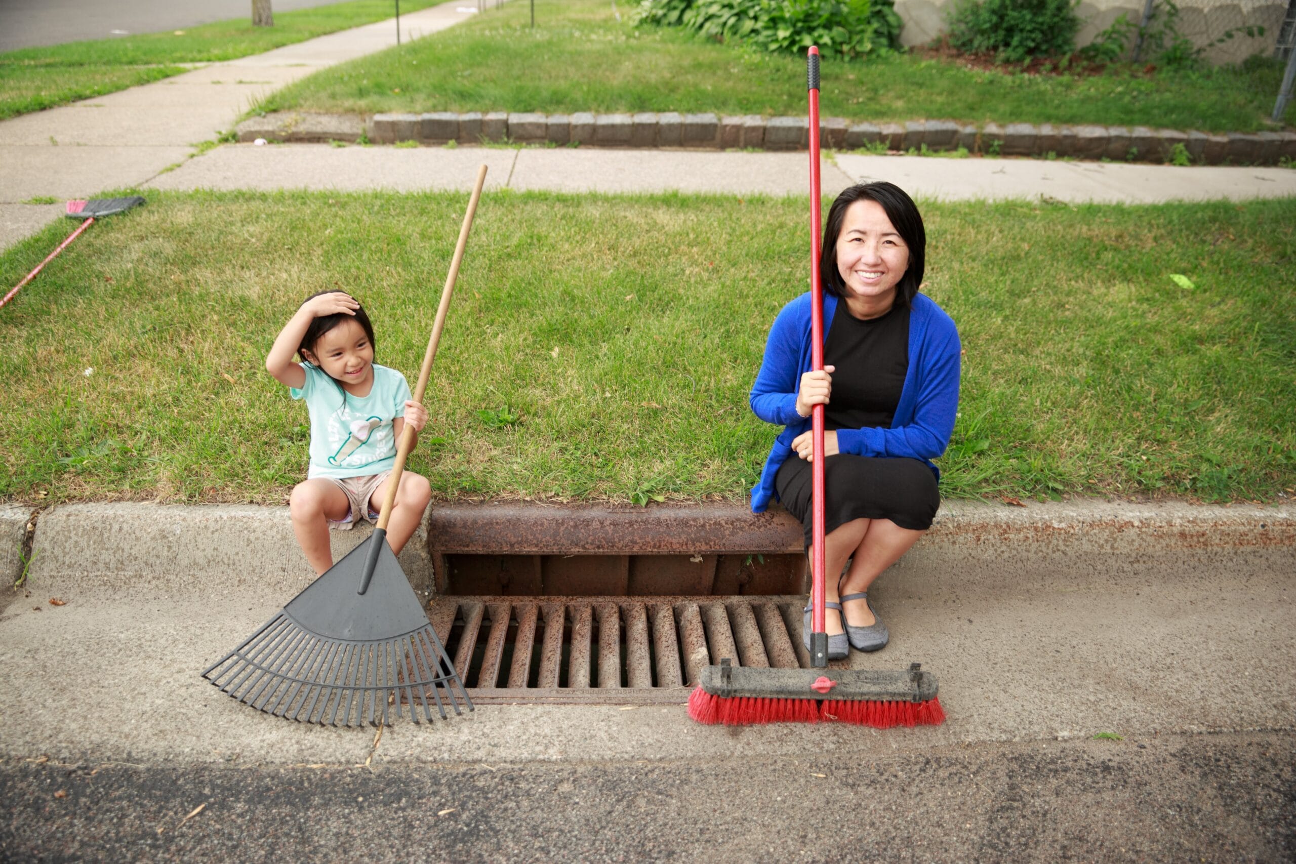 A adult woman and a child sitting on the curb next to a stormdrain each holding a rack